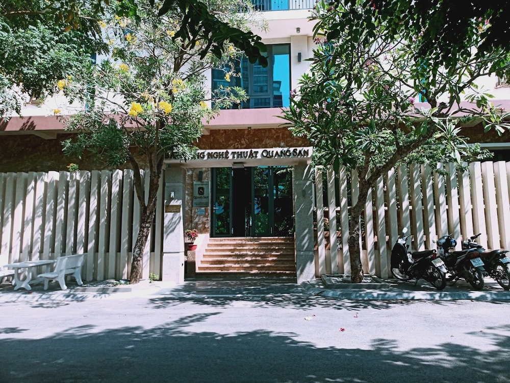 The modest exterior, with two lush trees by the entrance and a simple stone bench, offers a peaceful yet artistic ambiance